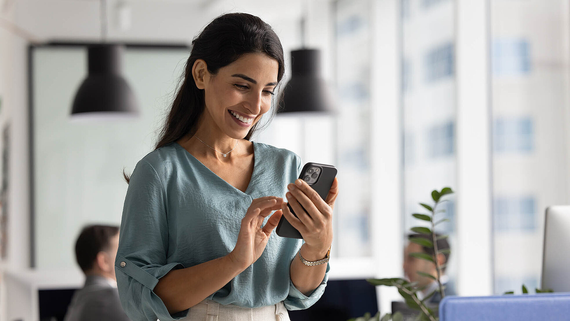 © fizkes - Adobestock.com Geschäftsfrau im modernen Büro lächelt beim Blick auf ihr Smartphone.