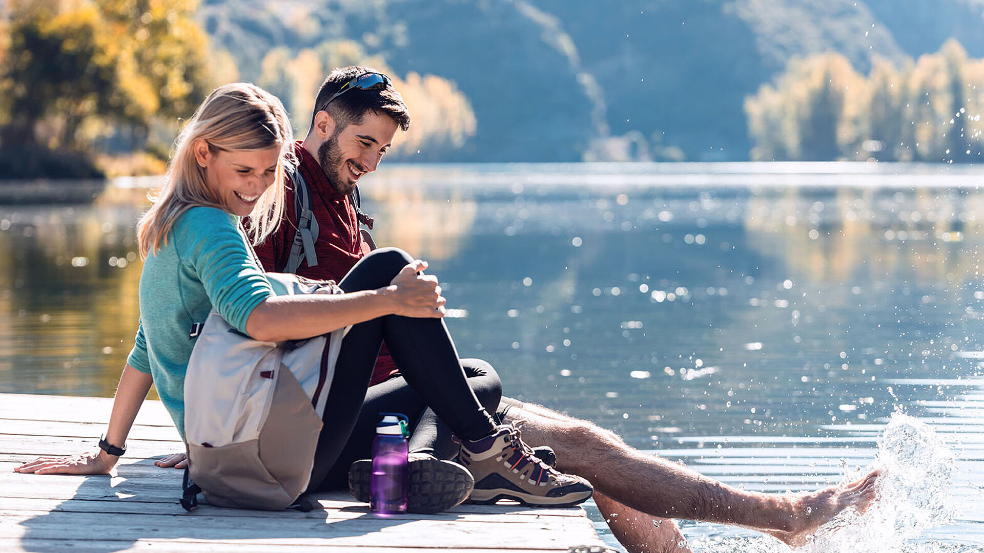 © nenetus - AdobeStock.com Lächelndes Paar sitzt mit Wanderrucksack auf einem Steg und spielt mit den Füßen im klaren Wasser eines Bergsees