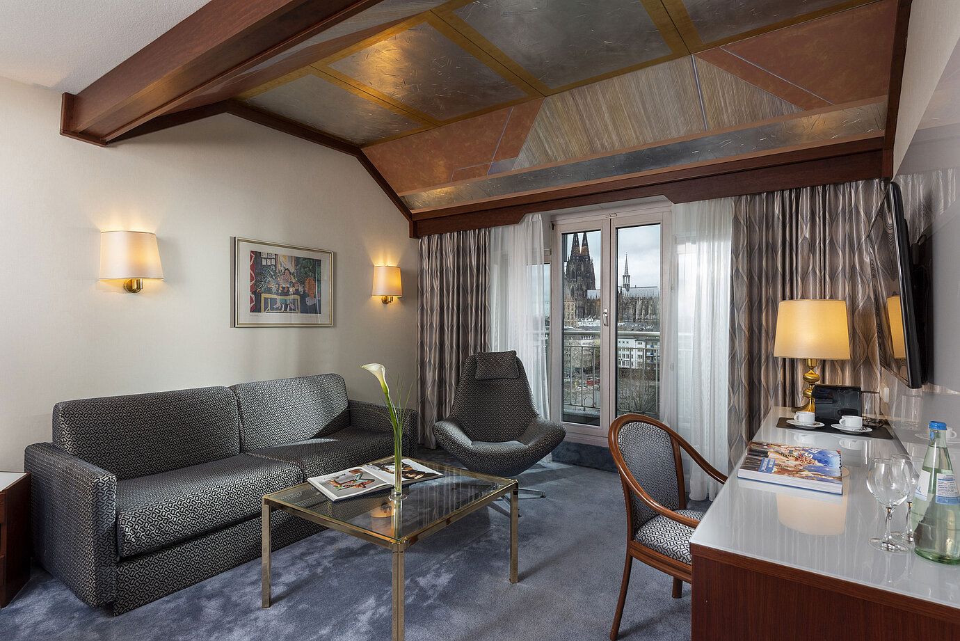 Junior Suite im Maritim Hotel Köln mit Blick auf den Kölner Dom, stilvollem Sofa, Sessel und Schreibtisch mit Lesematerial.