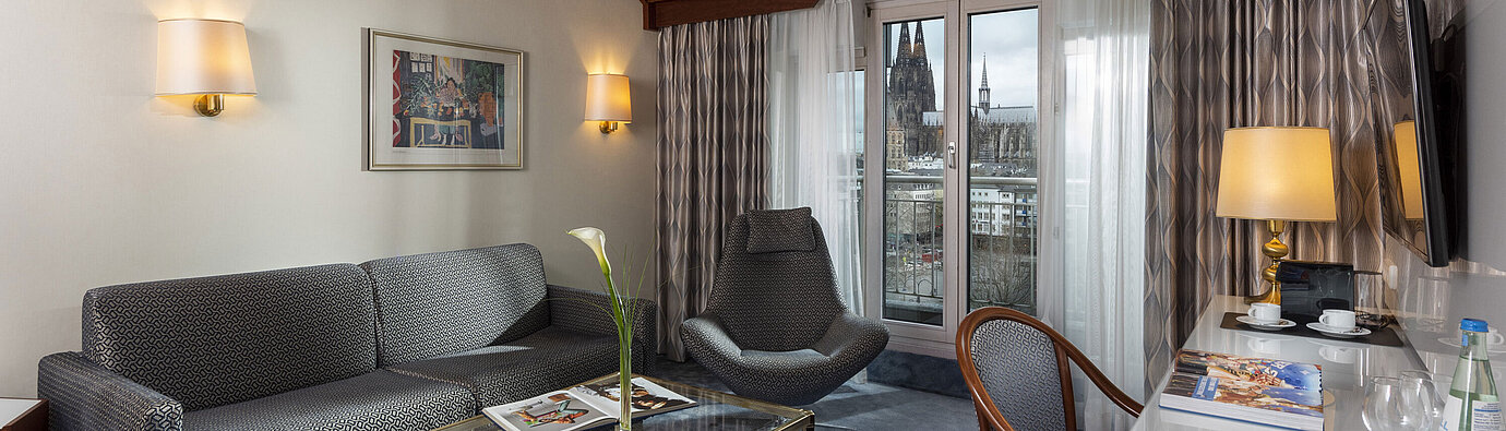 Junior Suite im Maritim Hotel Köln mit Blick auf den Kölner Dom, stilvollem Sofa, Sessel und Schreibtisch mit Lesematerial.
