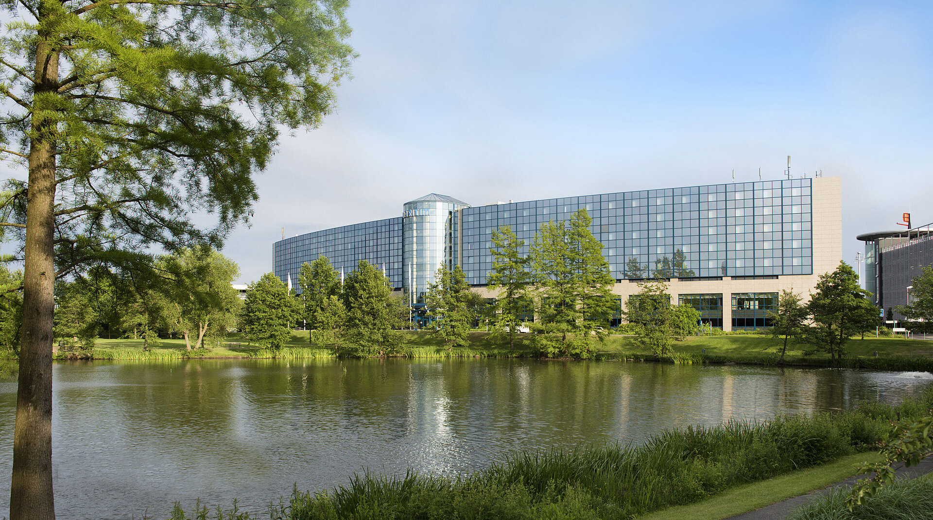 Außenansicht Das moderne Maritim Hotel Hannover mit Glasfassade und Blick auf den See inmitten einer grünen Landschaft.