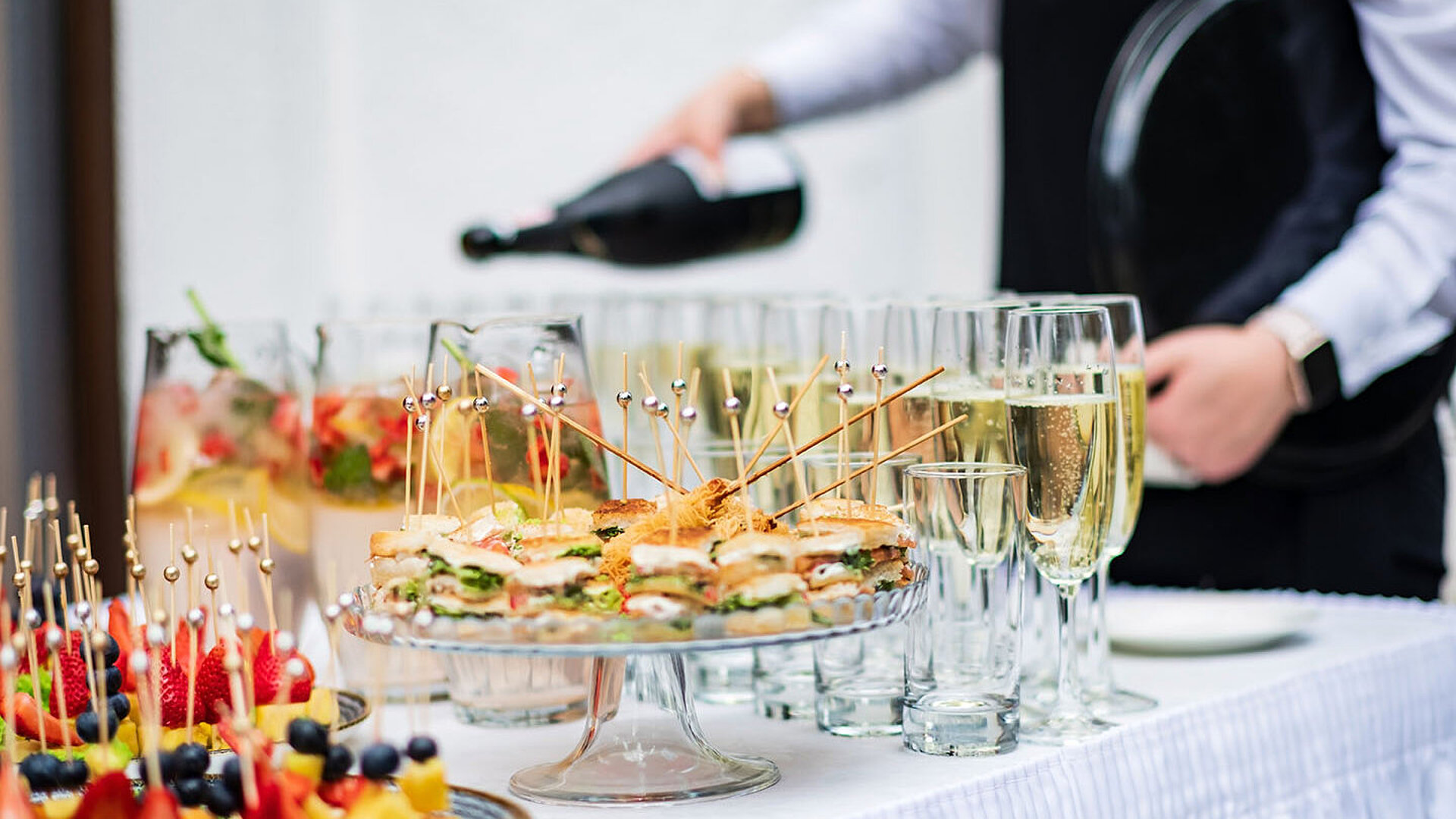 © Maksim Shebeko - AdobeStock.com Mitarbeiter schenkt Sekt für eine Veranstaltung ein, daneben stehen Sektgläser und Fingerfood bereit