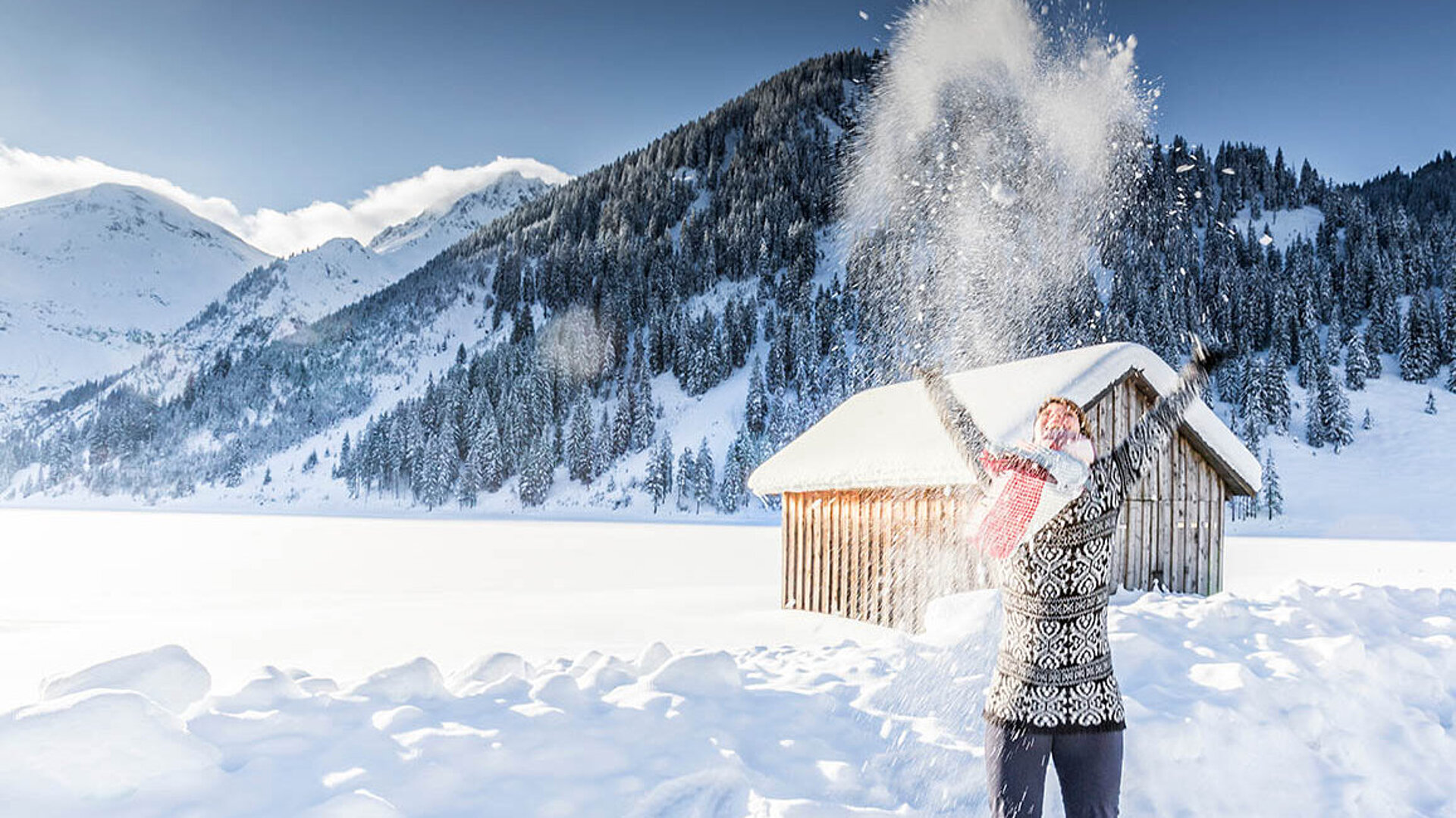 Person wirft Schnee in die Luft vor verschneiten Bergen und einer Hütte in strahlender Winterlandschaft.