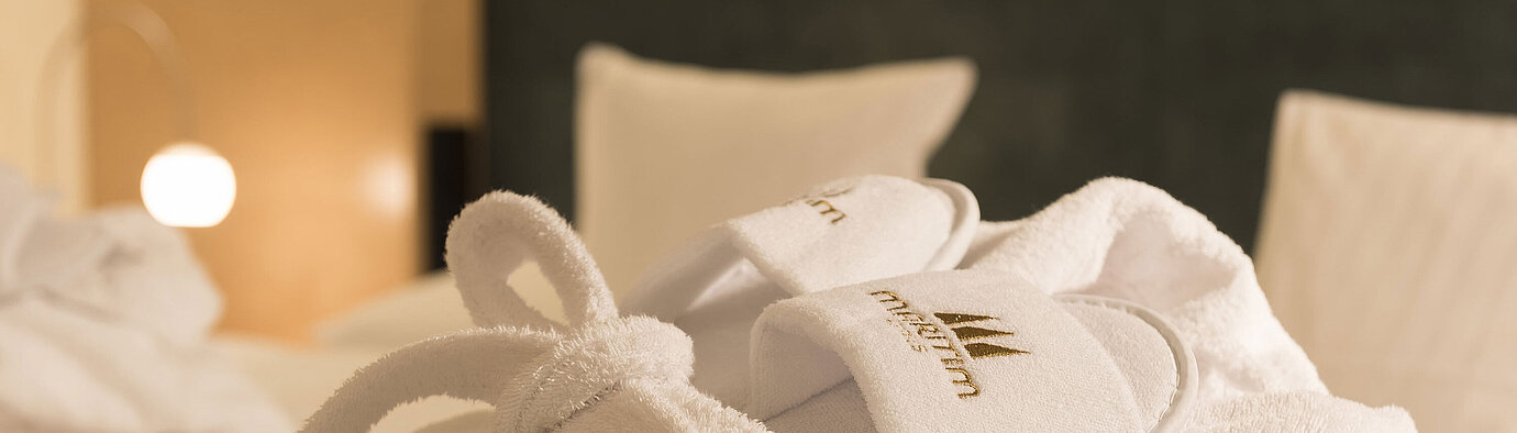 Ein flauschiger Bademantel und Hausschuhe mit Maritim-Logo auf einem Bett im Maritim proArte Hotel Berlin.