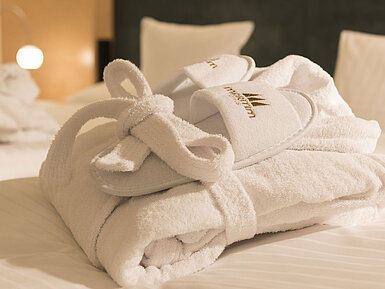 Ein flauschiger Bademantel und Hausschuhe mit Maritim-Logo auf einem Bett im Maritim proArte Hotel Berlin.