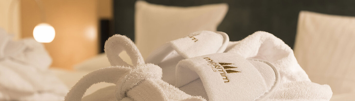 Ein flauschiger Bademantel und Hausschuhe mit Maritim-Logo auf einem Bett im Maritim proArte Hotel Berlin.