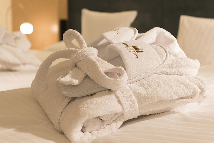 Ein flauschiger Bademantel und Hausschuhe mit Maritim-Logo auf einem Bett im Maritim proArte Hotel Berlin.