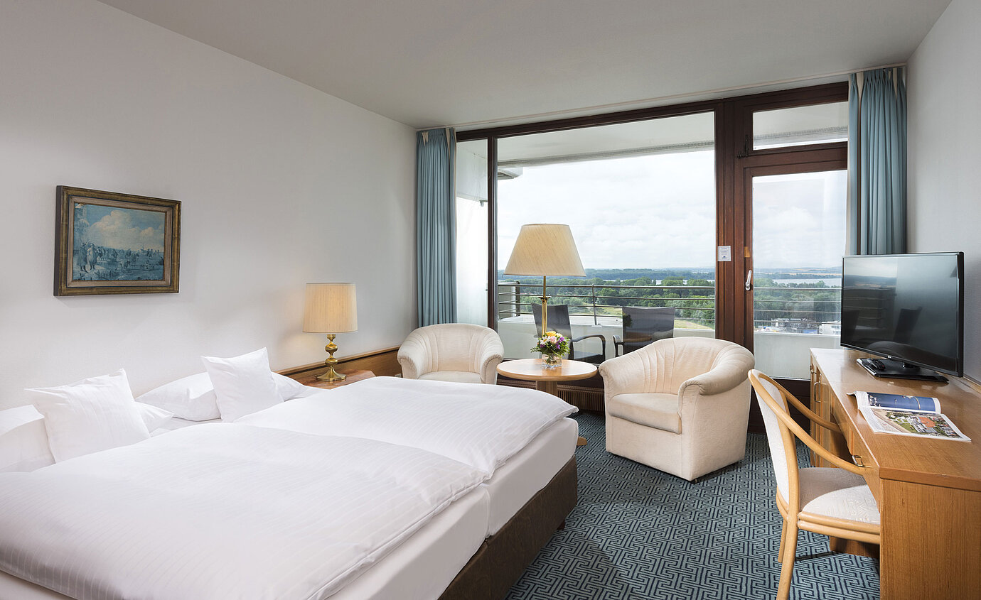 Doppelzimmer mit Sitzgruppe, Schreibtisch und Balkon mit Ausblick auf grüne Landschaft in Travemünde