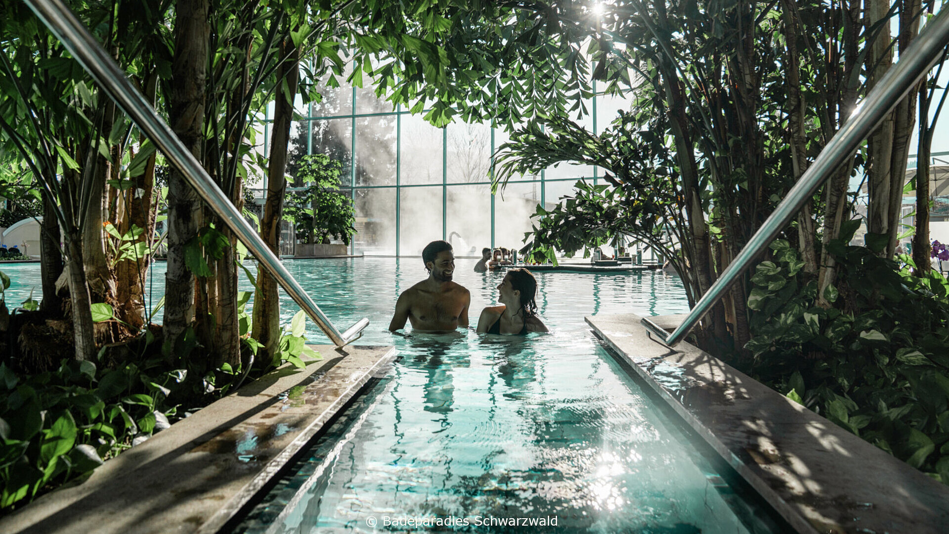 Zwei Personen entspannen im Indoor-Pool mit tropischer Begrünung und Tageslicht