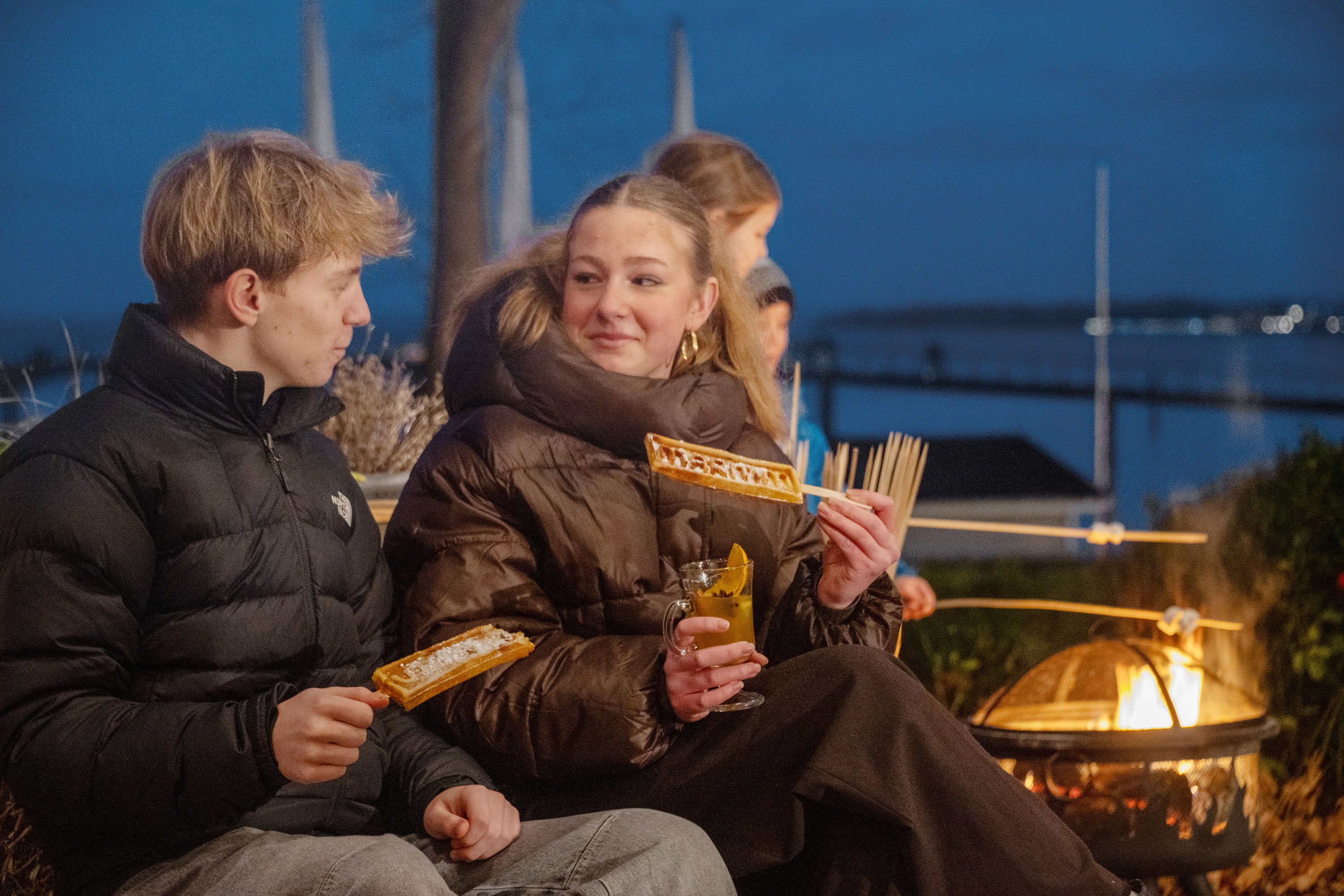 Winterzauber an der Ostsee Familie genießt frische Waffeln am Feuerkorb auf einer winterlich dekorierten Terrasse mit Ostseeblick.